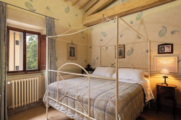 Una cama con dosel, un edredón estampado, un radiador, una ventana con cortinas y obras de arte colgadas en la pared.