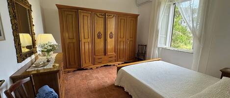 A bedroom with a wooden wardrobe, a bed with a white quilt, a chair with towels, a mirror, and a window with curtains.