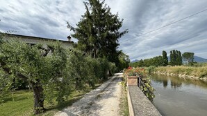 A narrow path beside a calm river, flanked by dense greenery and a building with a chimney.