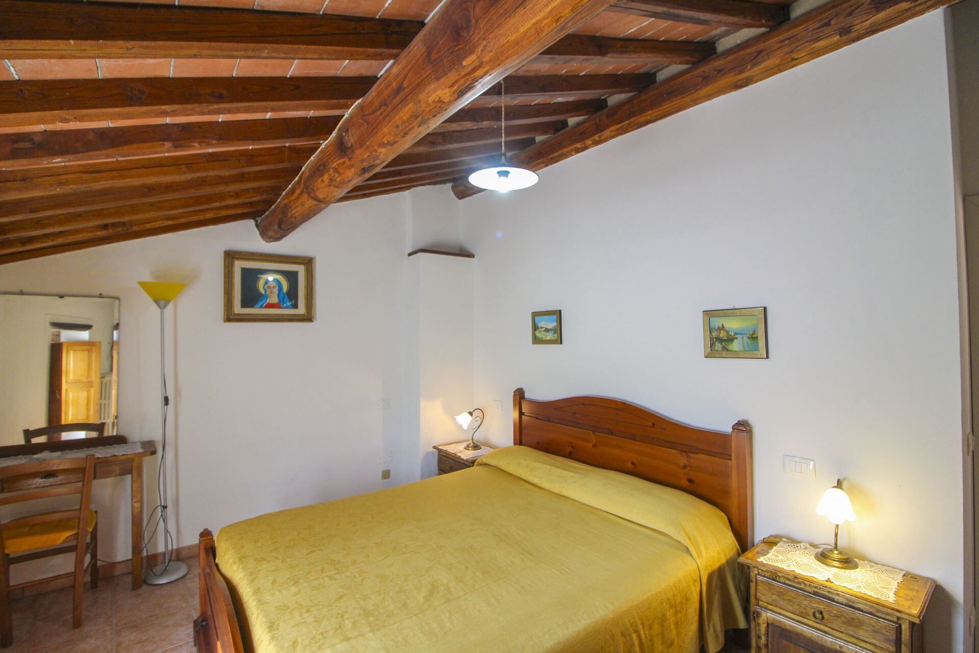 Una habitación con techo de madera, una cama con colcha amarilla, una mesita de noche con una lámpara y un escritorio con una silla.