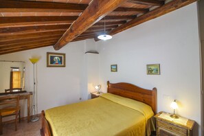 A room with a wooden ceiling, a bed with a yellow bedspread, a nightstand with a lamp, and a desk with a chair.