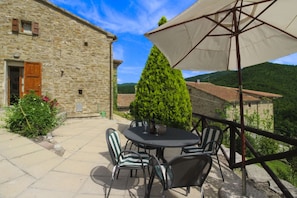 A stone building with a wooden door and window shutters, a round table with chairs, and a large umbrella. - Nice apartment for 4 guests with private pool, WIFI, TV and pets allowed (Castiglion Fiorentino)