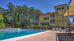 Ein Schwimmbecken mit gefliestem Deck, ein gelbes Gebäude mit grünen Fensterläden und umliegende Bäume.