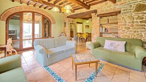 Una sala de estar acogedora con un sofá a cuadros, un sofá verde y un suelo de baldosas. Se observan vigas de madera en el techo y una pared de piedra con estanterías.