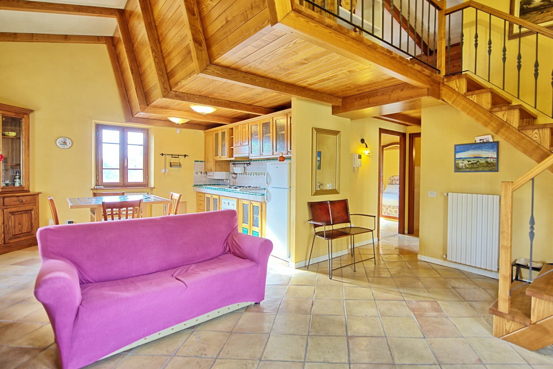 A spacious living area with a wooden staircase, a kitchen with wooden cabinets, and a dining area with a pink sofa.
