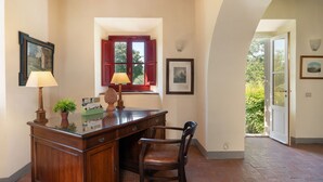 A room with a wooden desk, two lamps, a chair, a window with red shutters, and framed pictures on the walls.