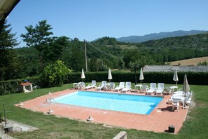 A rectangular swimming pool with a tiled deck, surrounded by white lounge chairs and umbrellas, set against a backdrop of lush greenery and distant hills. - Wonderful private villa for 14 guests with WIFI, private pool, TV, terrace and panoramic view (La Ginestra)