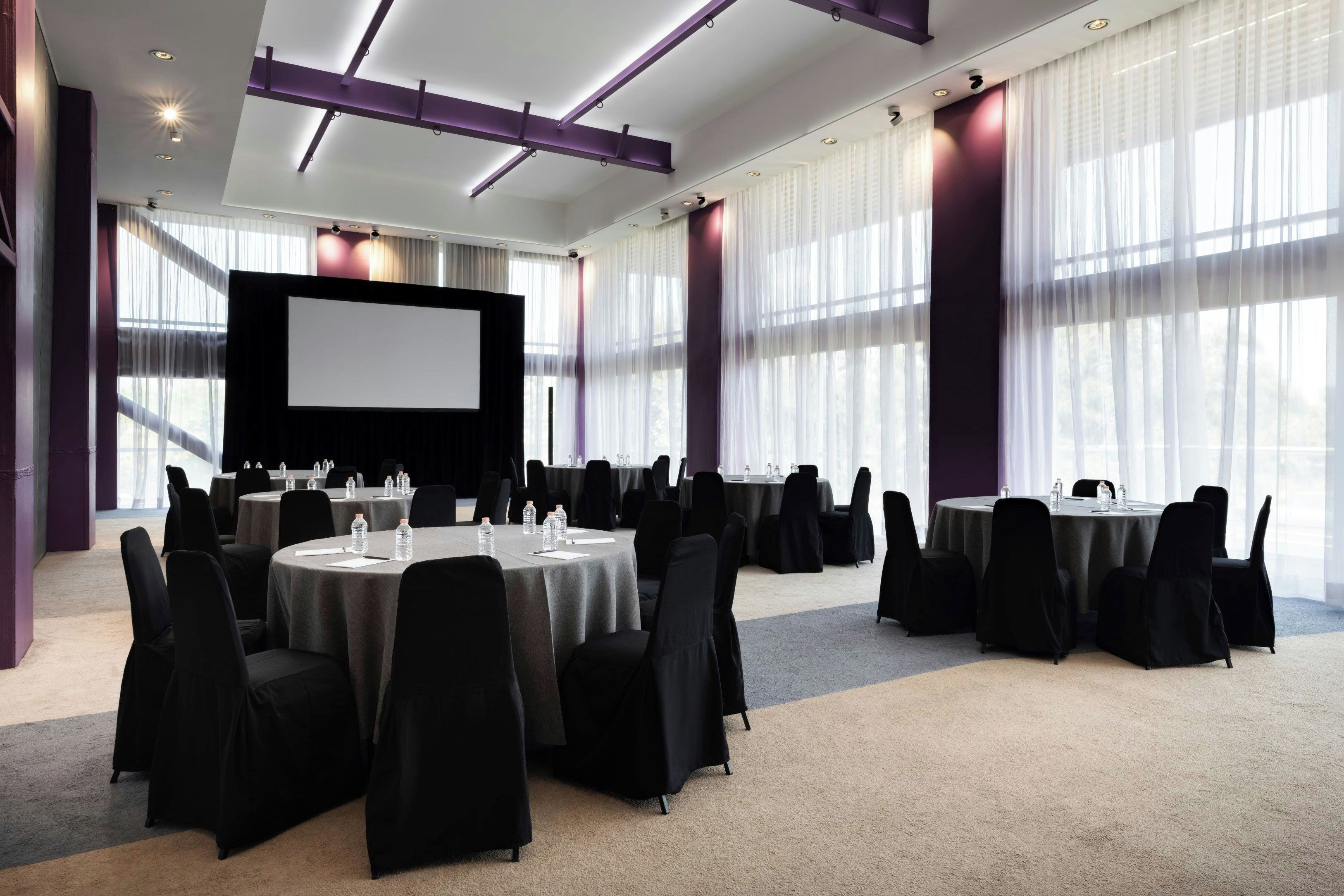 Een vergaderzaal met ronde tafels, stoelen, een groot scherm en een podium.