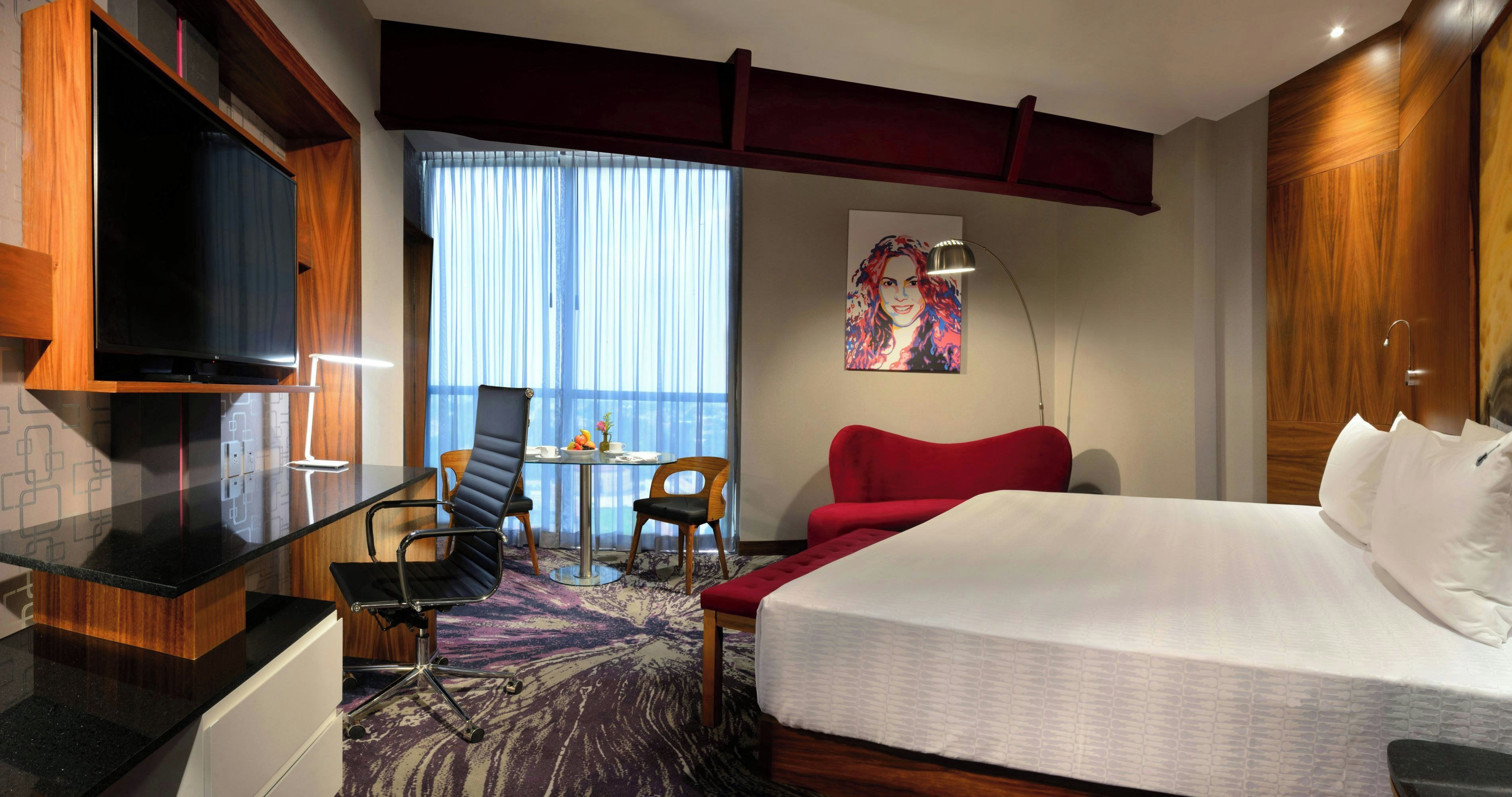 A hotel room with a bed, a desk with a chair, a television, and a window with curtains.