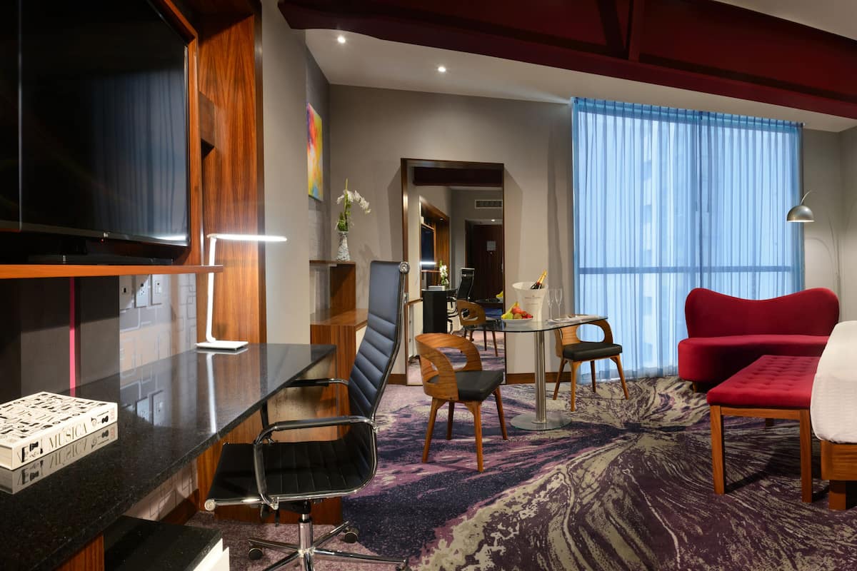 Ein Hotelzimmer mit einem Bett, einem Schreibtisch, einem Sessel, einem roten Sofa, einem Fernseher und einem Fenster mit Vorhängen.
