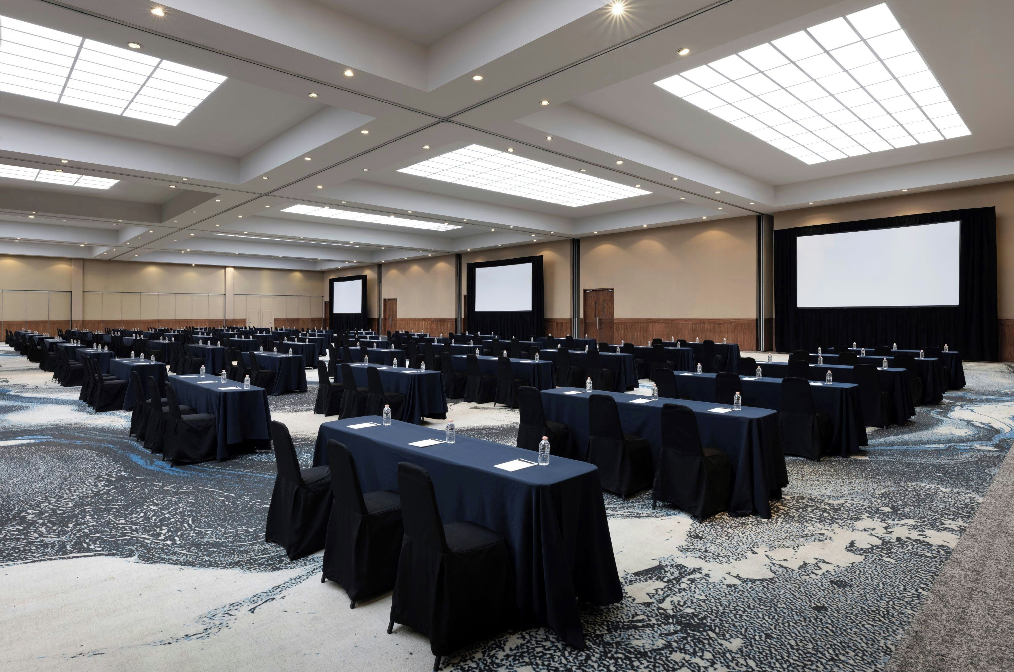 A conference room with rows of tables and chairs, large screens, and a carpeted floor.