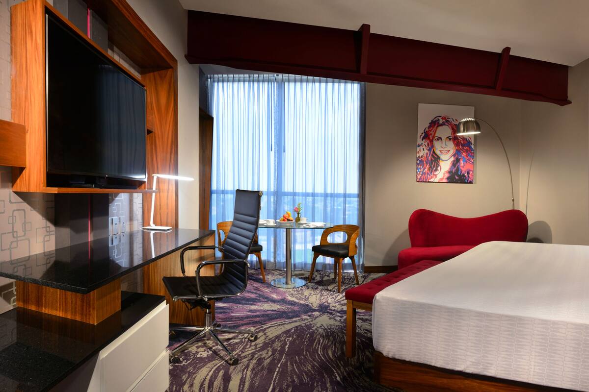 Ein Hotelzimmer mit einem Bett, einem Fernseher, einem Schreibtisch mit Stuhl, einem Tisch mit Stühlen und einem Fenster mit Vorhängen.