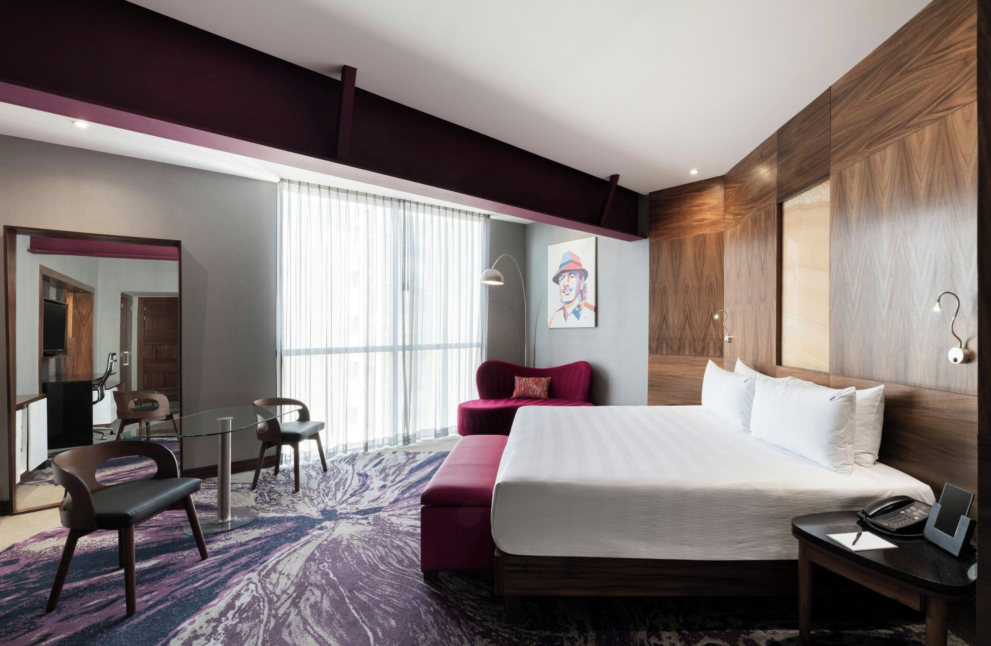 A modern hotel room with a large bed, a desk with a phone, a chair, a sofa, and a large window with sheer curtains.