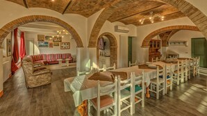 Zona pranzo con un lungo tavolo e sedie bianche, un divano e un televisore in una stanza con soffitti a volta in mattoni e pavimento in legno.