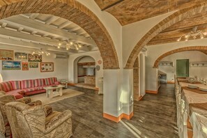 A spacious living area with a sofa, coffee table, and patterned armchairs. There are brick arches and a kitchen in the background.