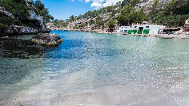 Paesaggio costiero con acque limpide, spiaggia sabbiosa e un edificio con porte verdi.