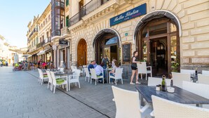 Un caffè all'aperto con tavoli e sedie, un cartello con la scritta Bella Italia e una strada acciottolata.