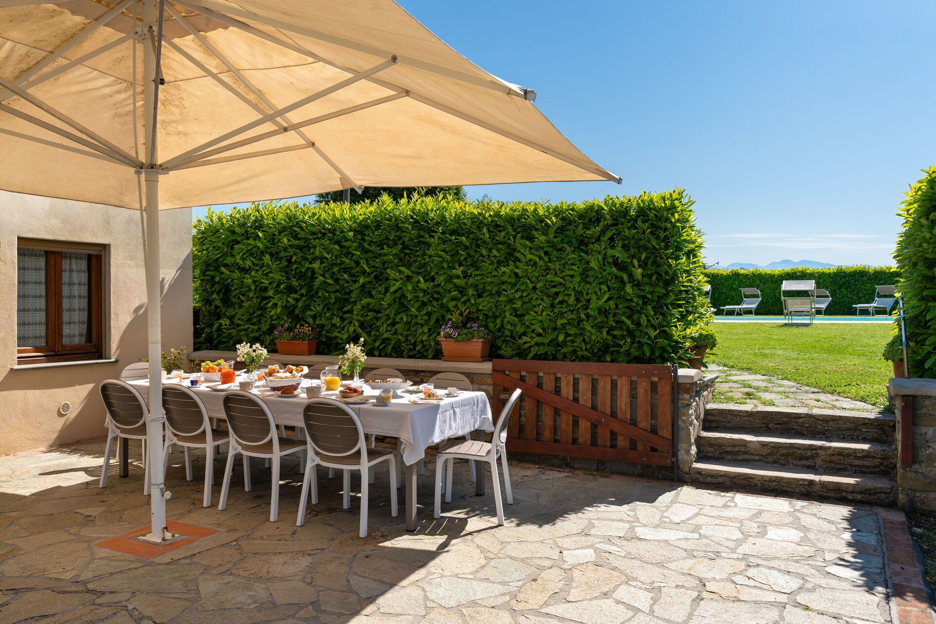 An outdoor dining area with a table set for six, surrounded by chairs, under a large umbrella.