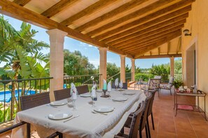 An outdoor dining area with a covered patio, wooden beams, and a view of a swimming pool and greenery.