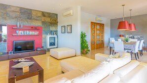 A modern living room with a flat-screen TV, a red wall-mounted shelf with books, a stone wall, and a dining area with a wooden table and chairs. - CAN BOU -  Villa with private pool only 4 km from the beach on the outskirts of Ses saline (Ses Salines)