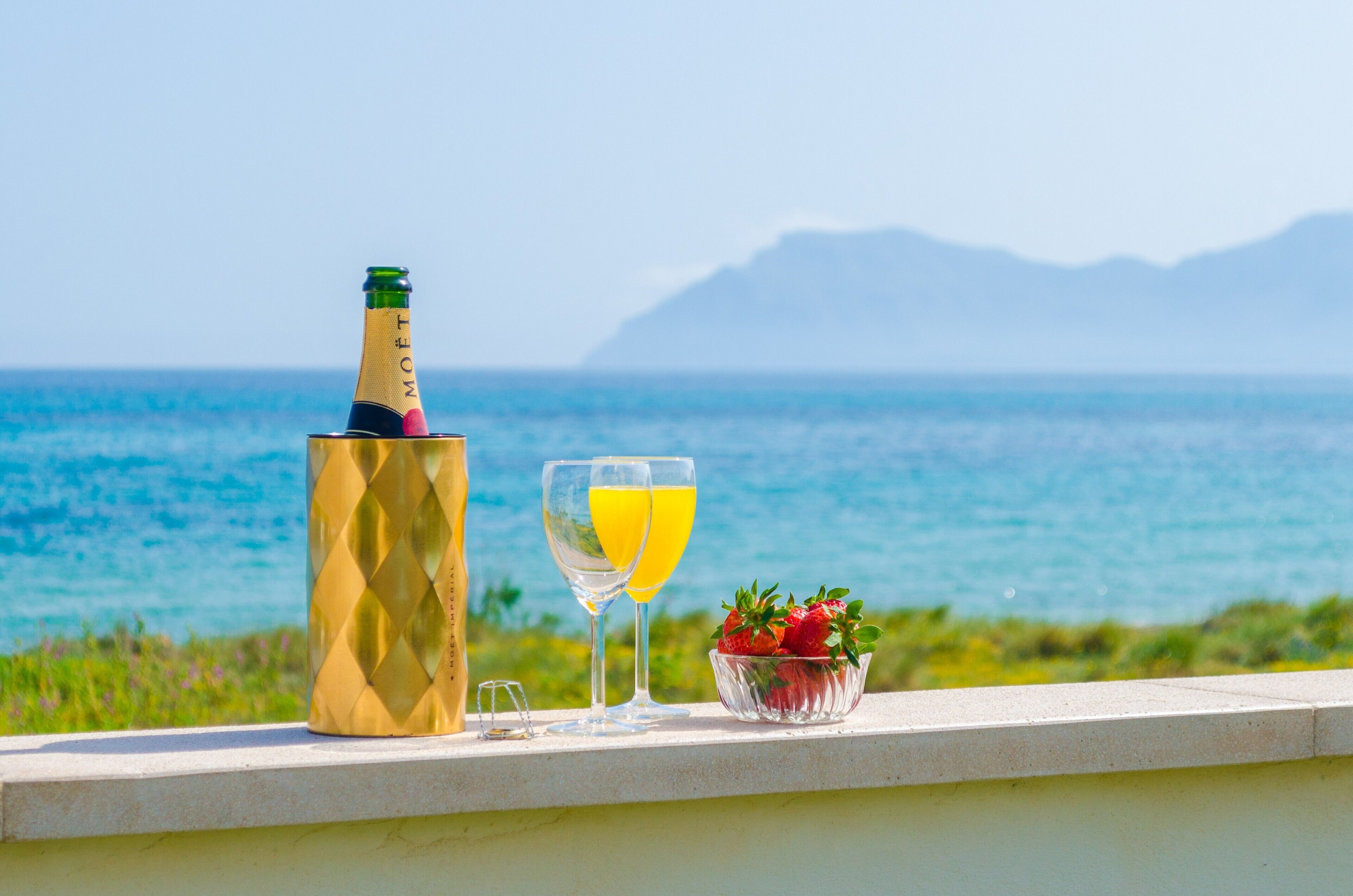 A bottle of champagne, a glass of orange juice, and a bowl of strawberries on a balcony with a view of the sea and mountains.