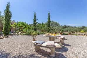 A garden patio with two stone benches, a table with fruits and a bowl, surrounded by potted plants and trees. - CAN PINA - ECO PINA - Apartment with shared pool in Costitx. Free WiFi (Costitx)