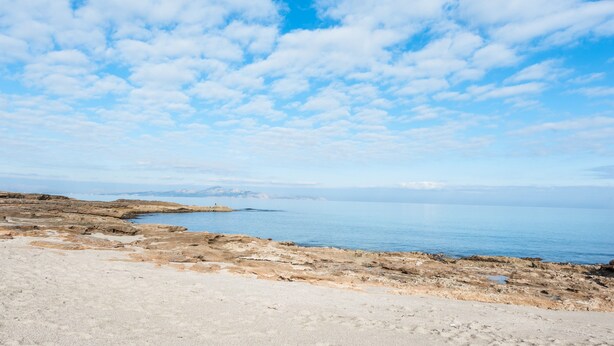 A coastal landscape with a rocky shoreline, a clear sky, and distant mountains.