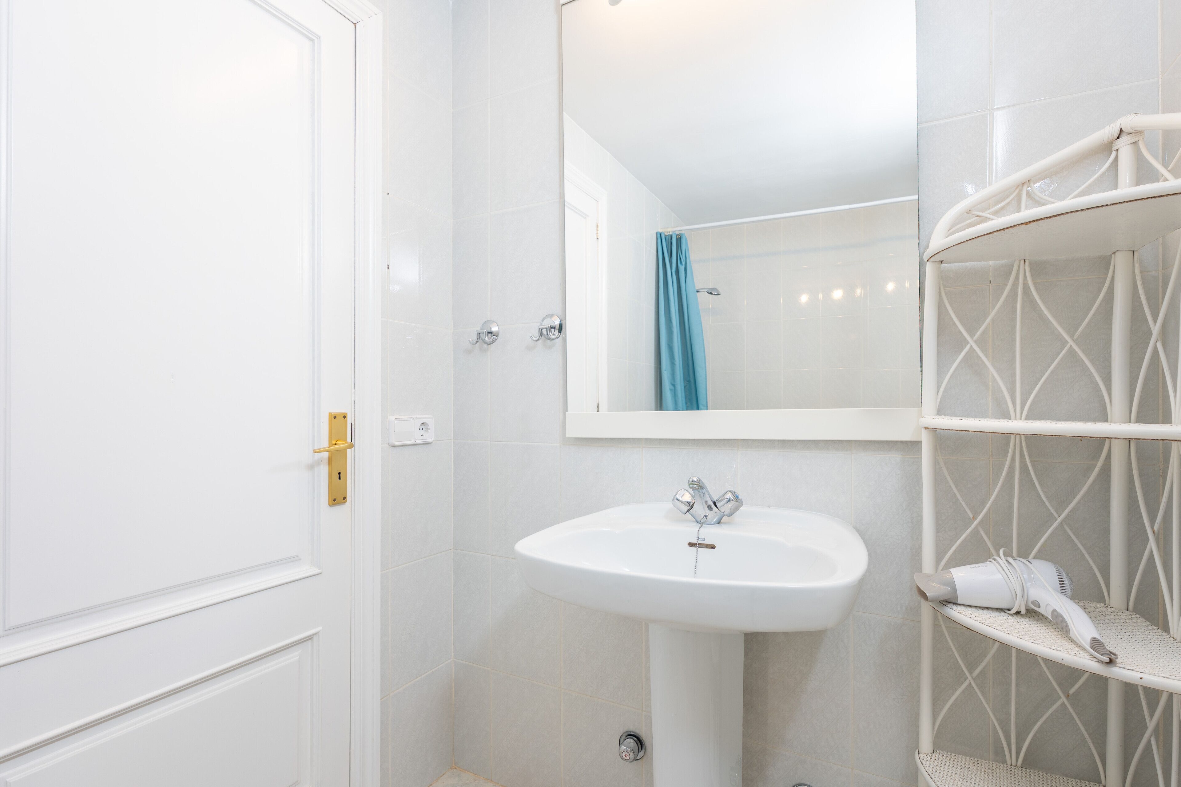 A bathroom with a white sink, a mirror, a shower curtain, and a white shelf with a hairdryer.