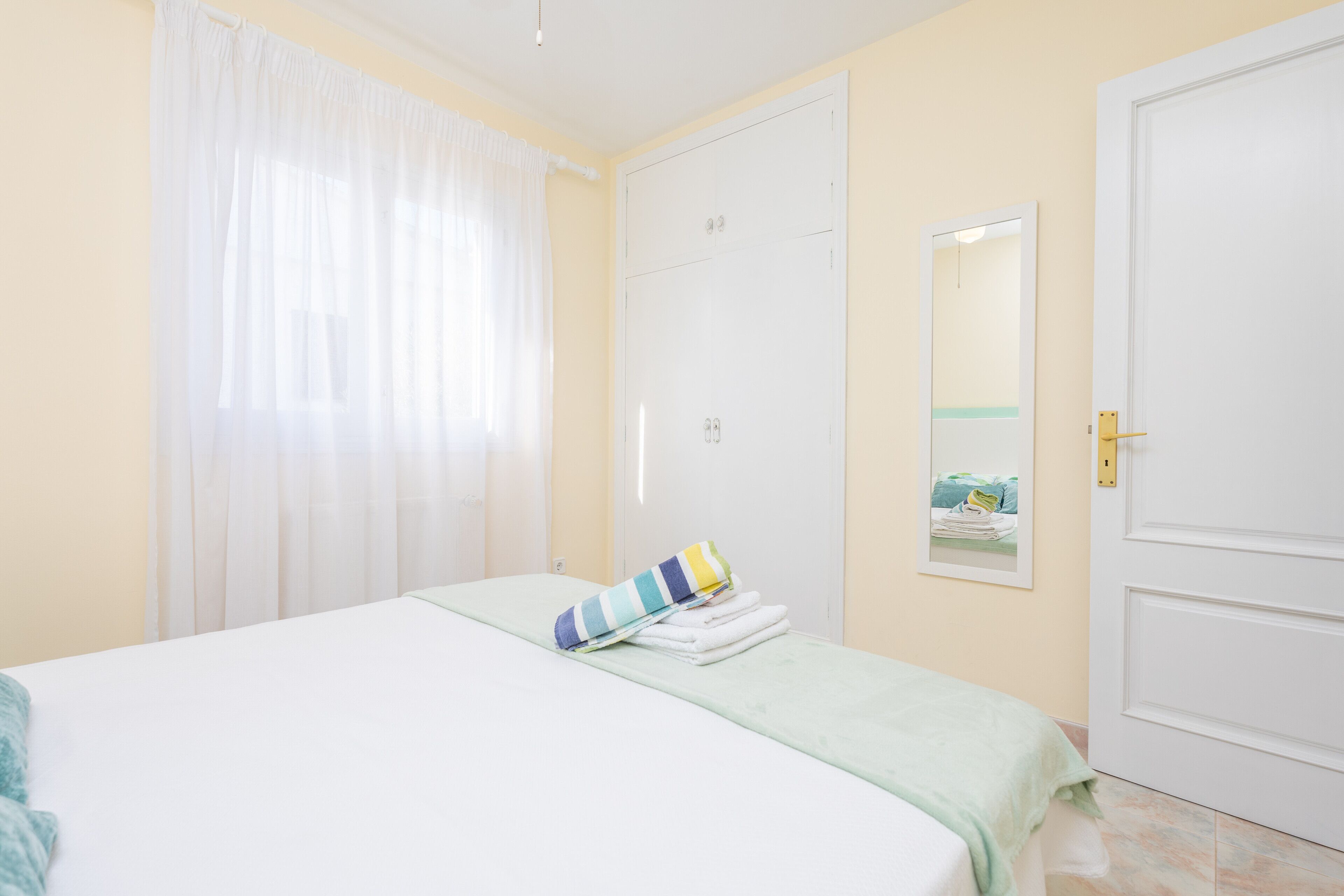 Un dormitorio con cama, una puerta blanca, un espejo y una ventana con cortinas.