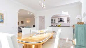 A modern dining area with a glass-top table, white chairs, and a wicker base. A kitchen is visible in the background with white cabinets and a green cabinet.