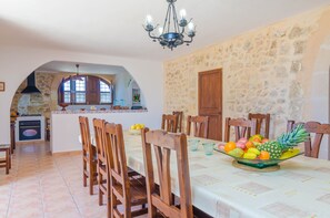 A dining area with a stone wall, wooden chairs, a table with a fruit bowl, and a kitchen in the background.