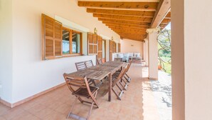Un espace extérieur abrité, aménagé pour les repas, avec une longue table et des chaises en bois, des poutres en bois au plafond et des fenêtres équipées de volets.