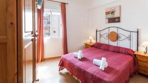 A hotel room with a bed, a wooden wardrobe, a window with curtains, a bedside table with a lamp, and a wall with a painting. - ODISEA - Apartment with sea views in Playa de Piles. (Playa de Piles)