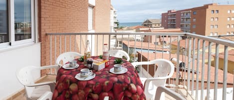 A balcony with a table set for two, white plastic chairs, and a red tablecloth with apple prints.