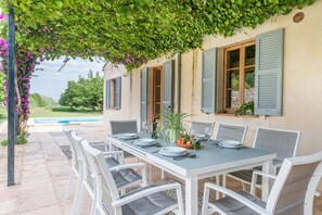 An outdoor dining area with a table set for six, surrounded by white chairs with grey cushions.