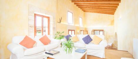 A spacious living room with a high wooden ceiling, white sofas with colorful cushions, a coffee table with books and a plant, and a stone wall with framed pictures.