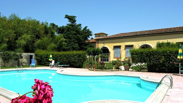 A swimming pool with a curved edge, surrounded by a paved area. There are lounge chairs and a small building with a tiled roof in the background.