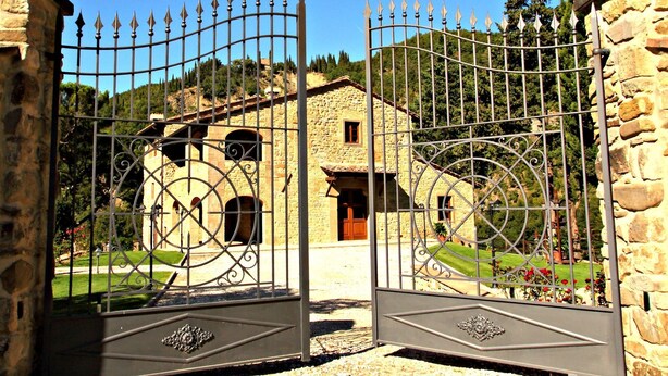 Un bâtiment en pierre à l’architecture traditionnelle, une porte en métal agrémentée d’éléments décoratifs et un jardin bien entretenu.