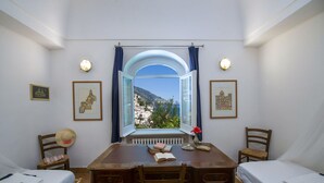 Ein Zimmer mit Schreibtisch, zwei Stühlen, einem Bett und einem Fenster mit Blick auf eine Küstenstadt.