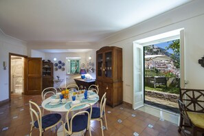 Un coin repas avec une table ronde dressée pour quatre personnes, entourée de chaises. Un buffet en bois avec des portes en verre se trouve à droite. La pièce offre une vue sur une colline avec des bâtiments.