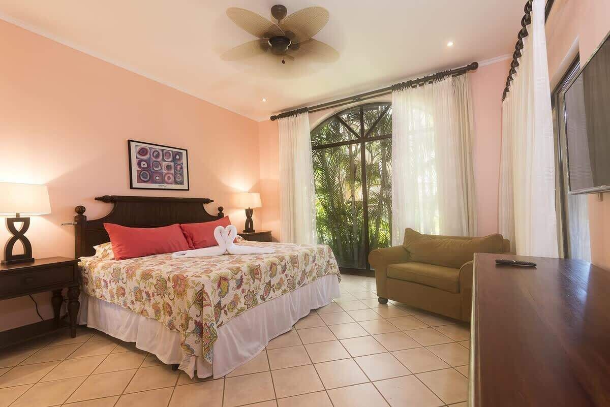 Un dormitorio con cama, mesita de noche, silla, ventilador de techo y ventana con cortinas.