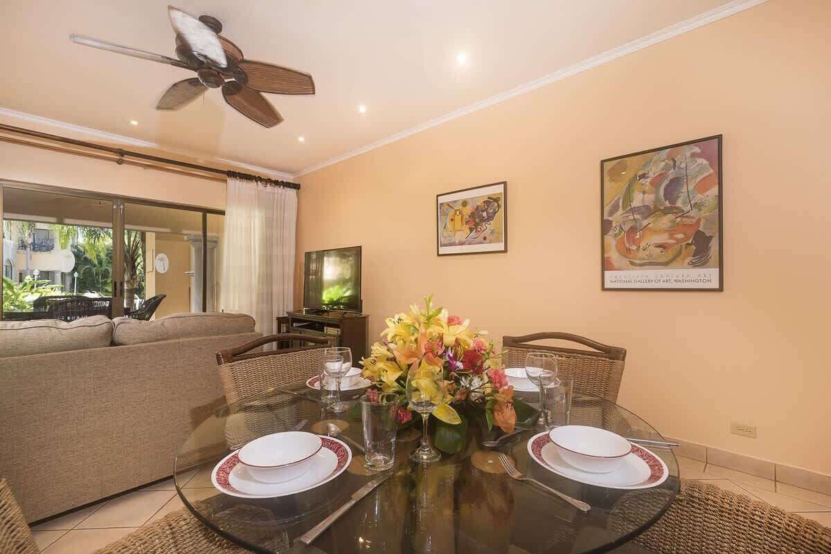 Un coin repas comprenant une table ronde en verre, deux chaises, un canapé, un ventilateur de plafond et des œuvres d’art accrochées aux murs.