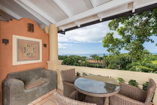 Un balcon agrémenté de meubles en osier, d’une table ronde en verre et offrant une vue sur l’océan et la végétation.
