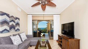 A living room with a sofa, coffee table, TV, and ceiling fan. - Sunrise 23- 2 Bedroom Ocean View Condo (Tamarindo)