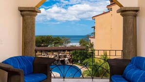 A balcony with two blue cushioned chairs, a small pool, and a view of the sea and buildings. - Sunrise 23- 2 Bedroom Ocean View Condo (Tamarindo)