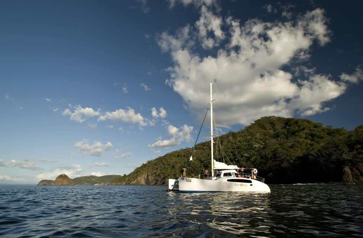 Un barco blanco con mástil y velas sobre aguas tranquilas, rodeado de colinas verdes exuberantes y un cielo despejado con nubes dispersas.