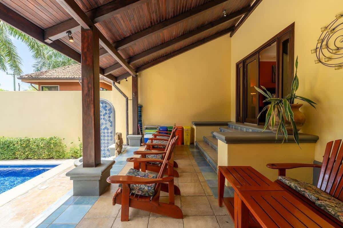Un área de estar al aire libre con techo, sillas de madera y piscina.