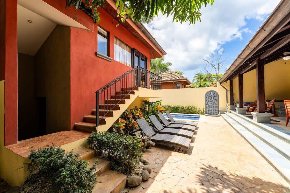 Una casa moderna con fachada roja, una escalera de piedra, piscina y mobiliario de exterior.