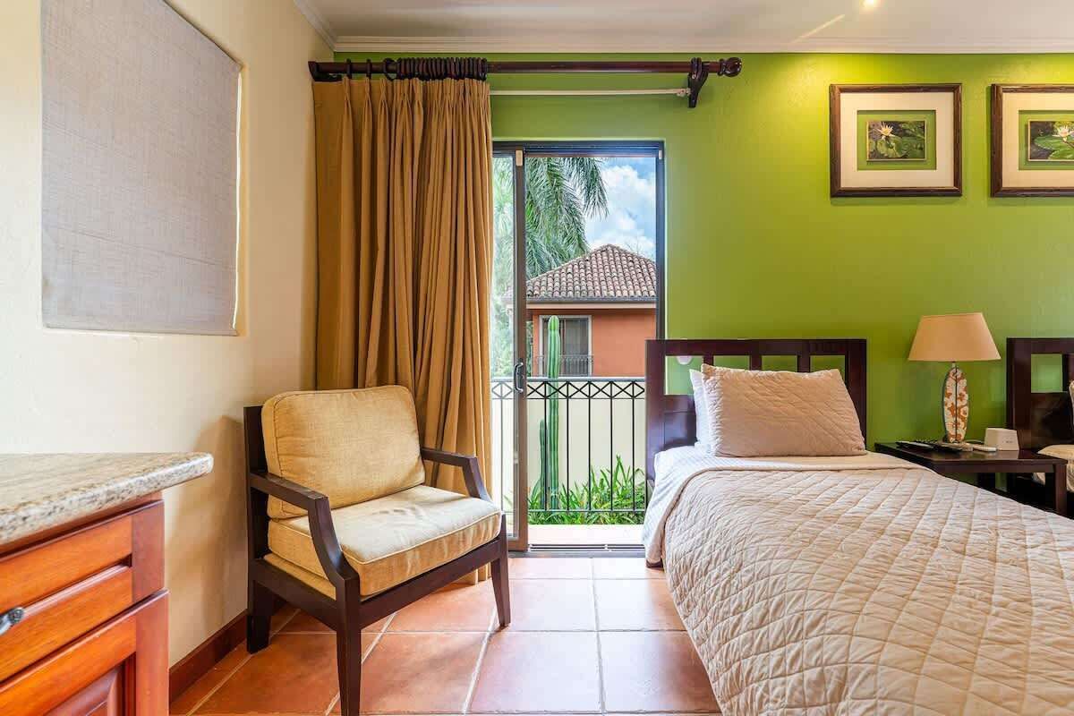 A hotel room with a bed, a chair, a nightstand, a lamp, and a view of the outside.