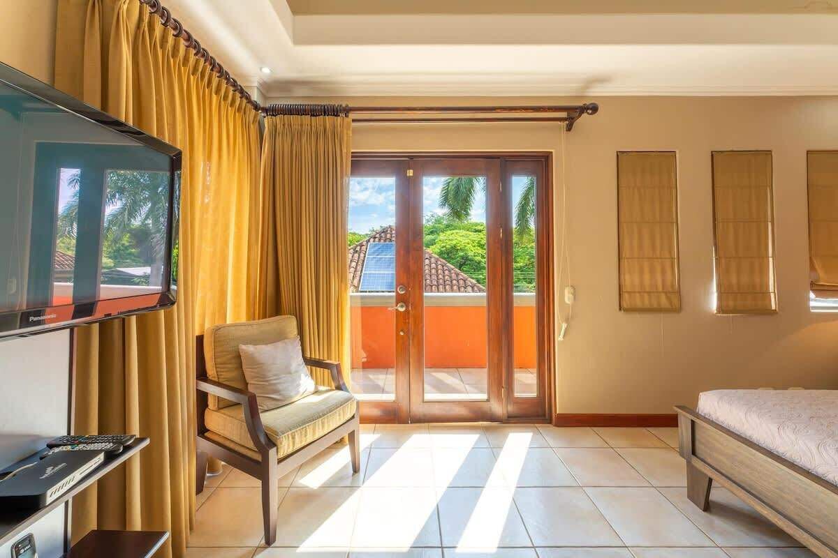 A hotel room with a television, a sofa, a bed, and a sliding glass door leading to a balcony with a view of palm trees and a building.